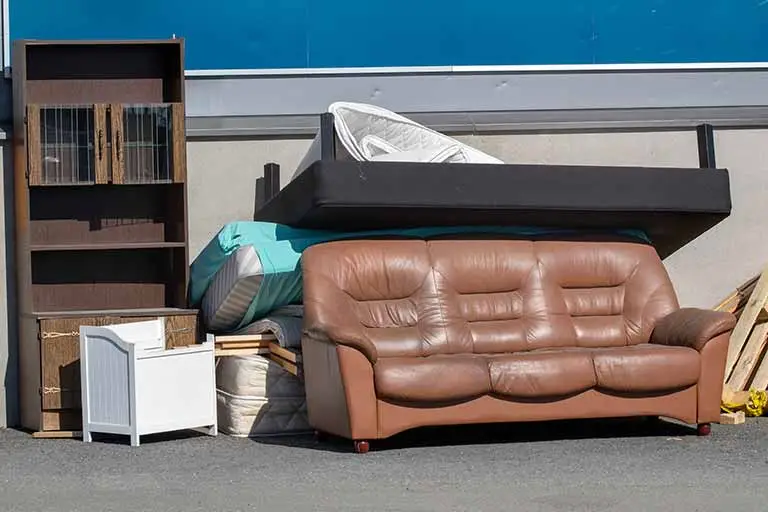 Furniture stacked for recycling, including a brown leather sofa, a mattress, a bookshelf, and a small white nightstand, highlighting the importance of responsible furniture disposal and recycling options.