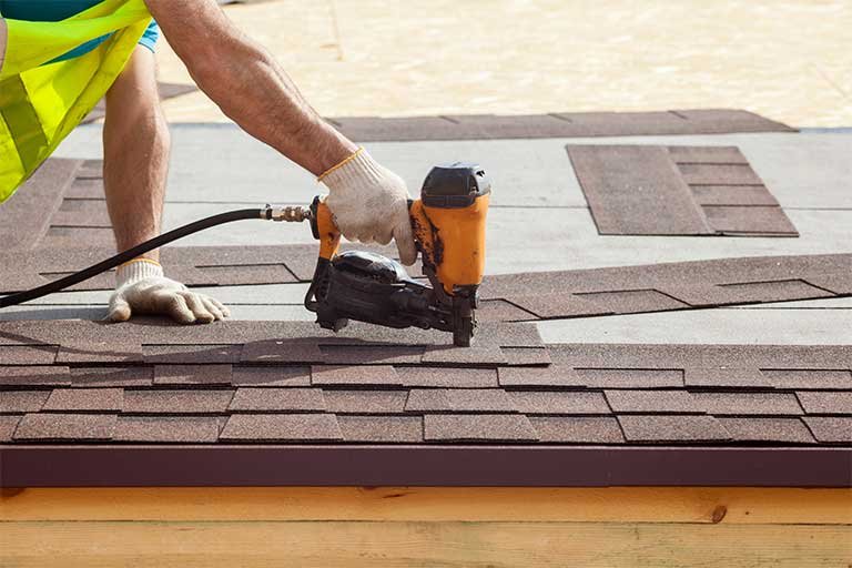 Roofer using a nail gun to install asphalt shingles on a roof, illustrating efficient roofing practices and the importance of proper waste management for debris disposal.
