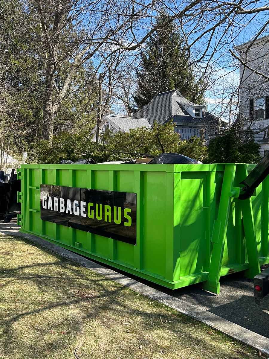 20 yard green dumpster with "GARBAGE GURUS" logo, positioned on residential street, surrounded by trees and houses, illustrating dumpster rental service for cleanouts and debris removal.